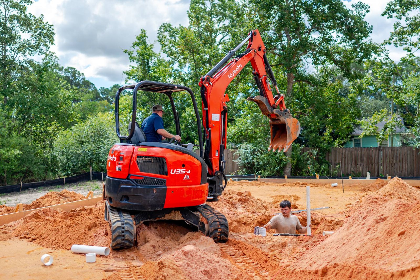 Excavator trenching while a Prestige Plumbing plumber sets underground drain lines